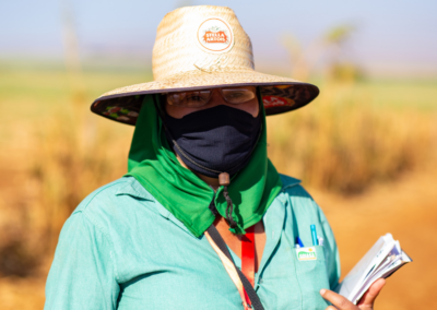 A woman wearing protective gear (a straw hat and mask over her mouth) and holding clipboard outside on a Fair Trade Certified Sugar Farm
