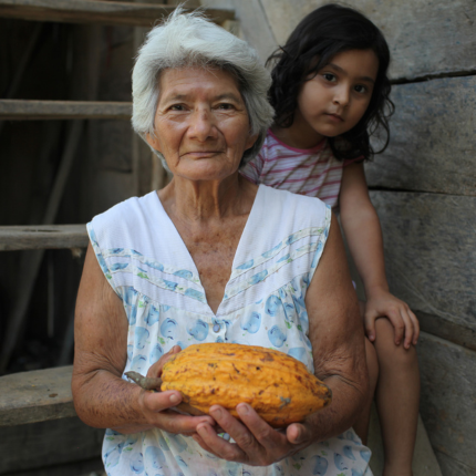 Elu Valdez, a life-long cacao grower, holds a cacao pod while sitting on her steps with her great-granddaughter